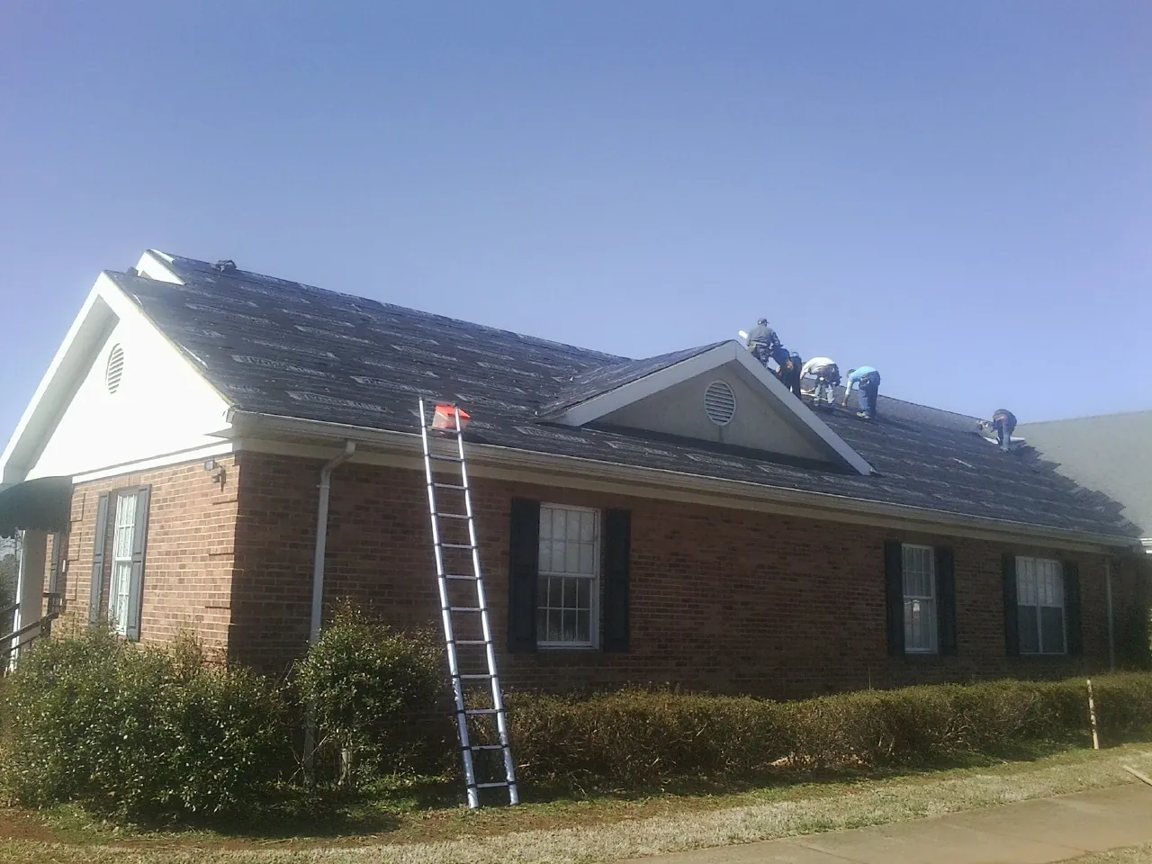 Crew tearing shingles off a brick ranch roof with a ladder set — Emergency Roof Repair work by our crew in Chanute