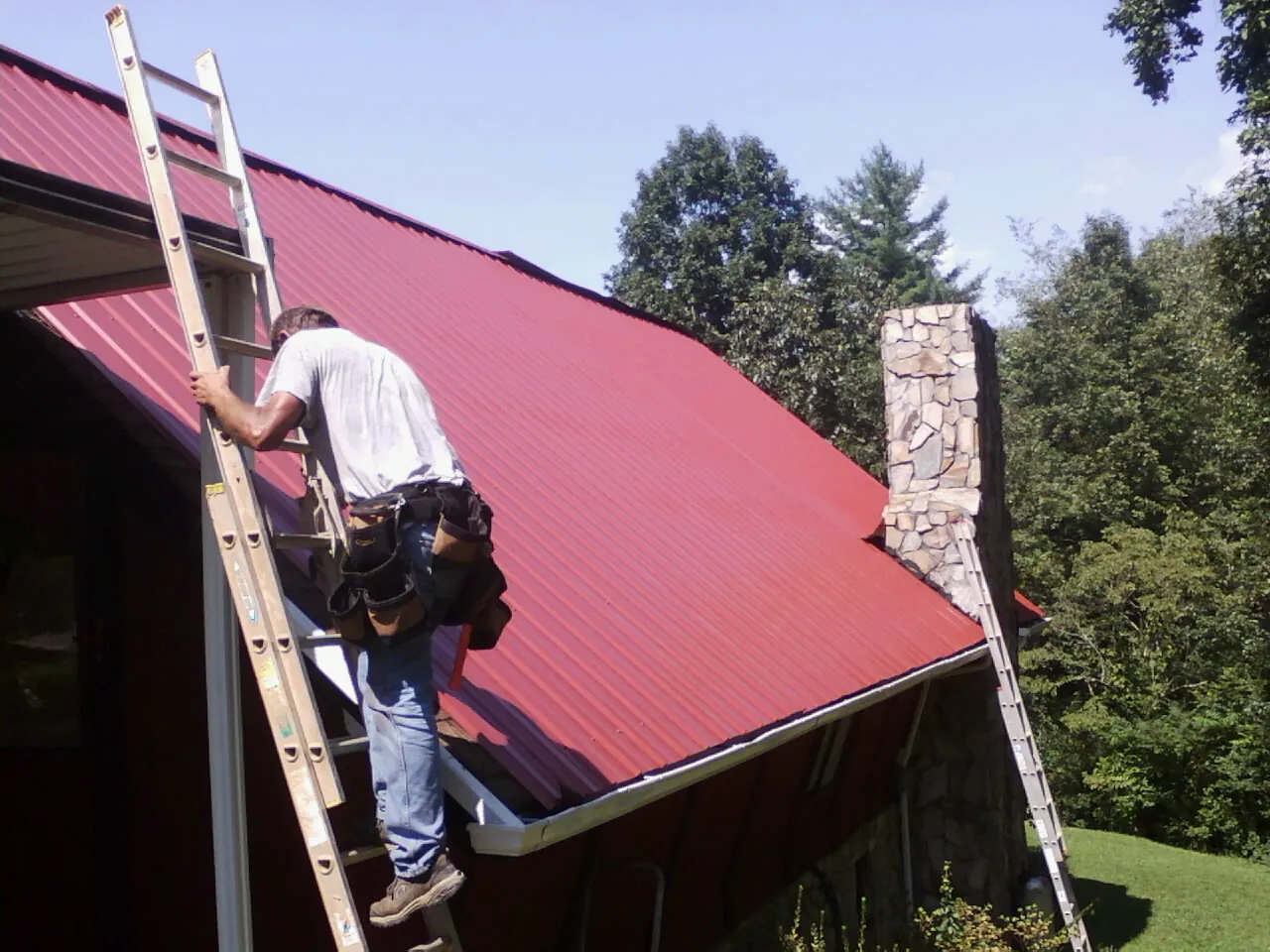 Roofer on a ladder beside a red metal roof and stone chimney — Soffit & Fascia Repair by Roofing Brothers in Chanute
